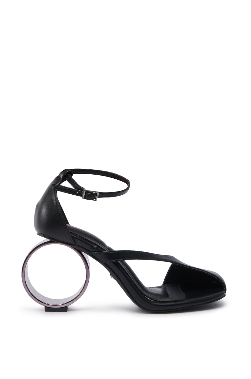 The AZALEA WANG CORABELLA BLACK NOVELTY HEEL SANDAL showcases a sculpted circular heel, ankle strap, and sleek faux leather upper, all set against a plain white background.