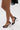 A person wearing the AZALEA WANG CORABELLA BLACK NOVELTY HEEL SANDAL with a large circular heel and ankle strap stands on a white surface; part of a black lace garment is also visible.