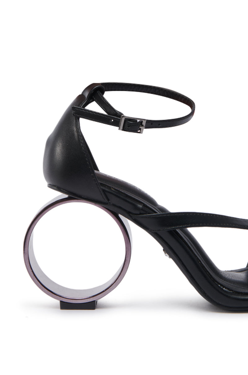 The AZALEA WANG CORABELLA BLACK NOVELTY HEEL SANDAL has a faux leather upper, ankle strap, and a sculpted heel with a circular hollow design for a bold, modern look.