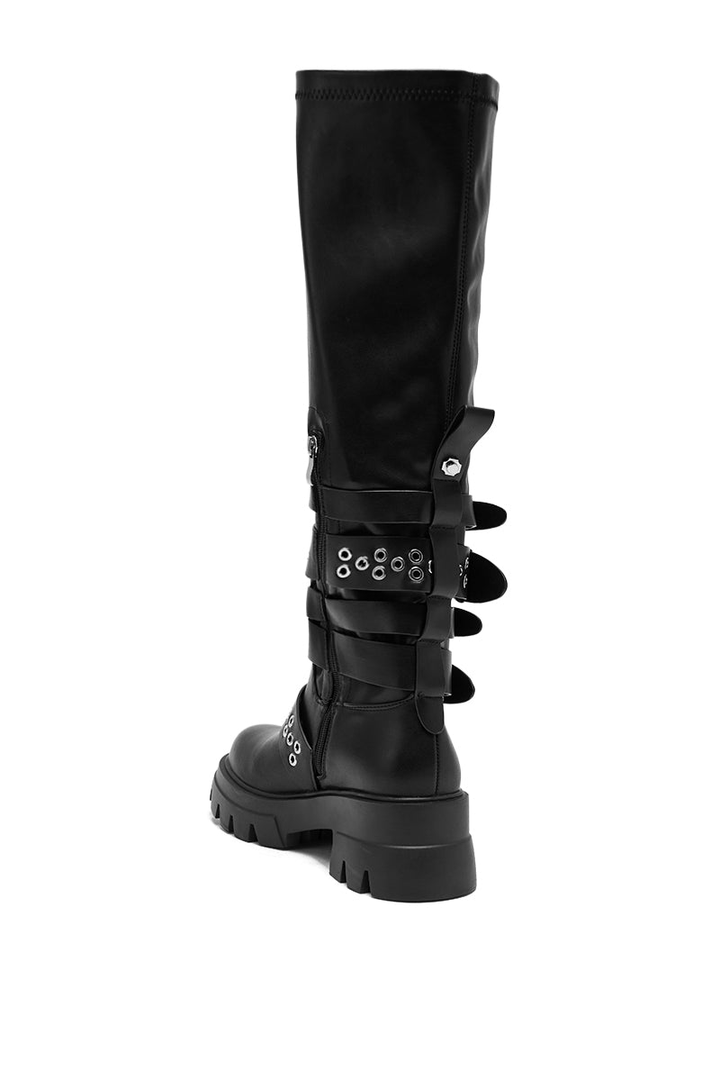 The AZALEA WANG CORDOVAN BLACK BELTED BOOT is a black faux leather knee-high boot with a chunky sole, decorative belted straps with metal eyelets, and silver-tone hardware, shown from the back and slightly to the side.