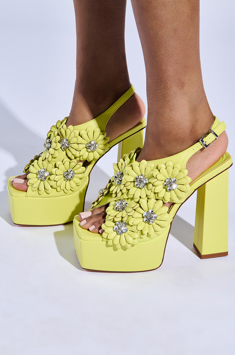A close-up shows someone wearing the AZALEA WANG CORRA lime platform sandals made from vegan leather, featuring large floral embellishments with jeweled centers, matched by high chunky heels and an open-toe design.