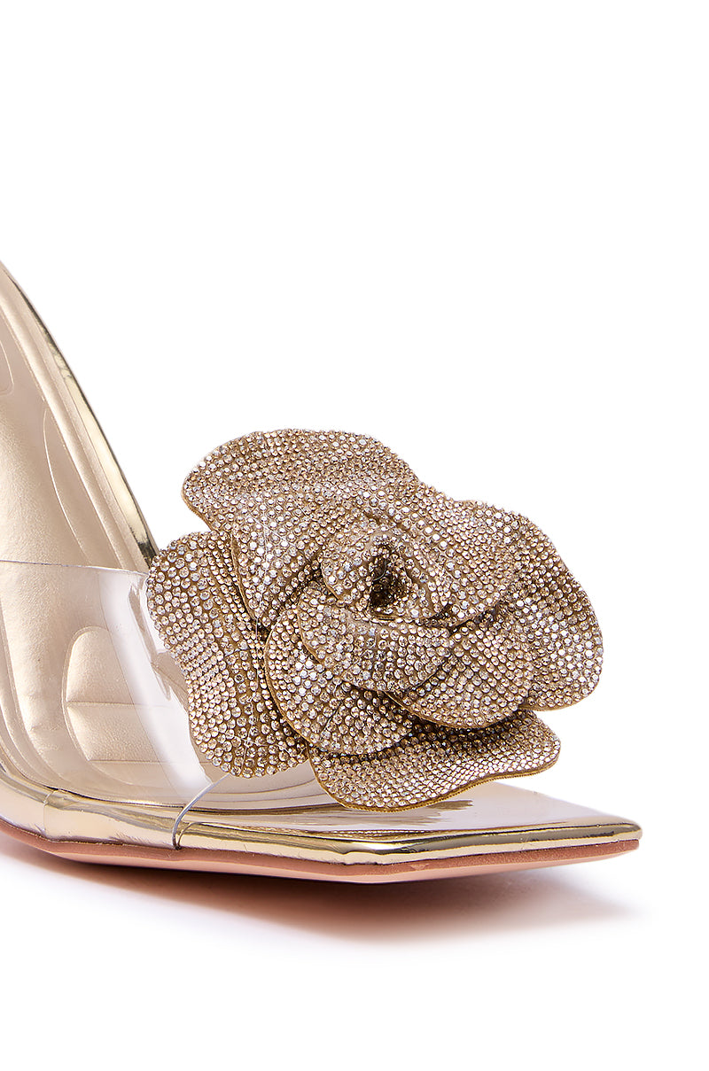 Close-up of the AZALEA WANG COSETTA GOLD PVC slip-on stiletto featuring a transparent side, sparkling gold flower rhinestone applique on the toe, shiny gold sole, and elegant gold heel design. 10