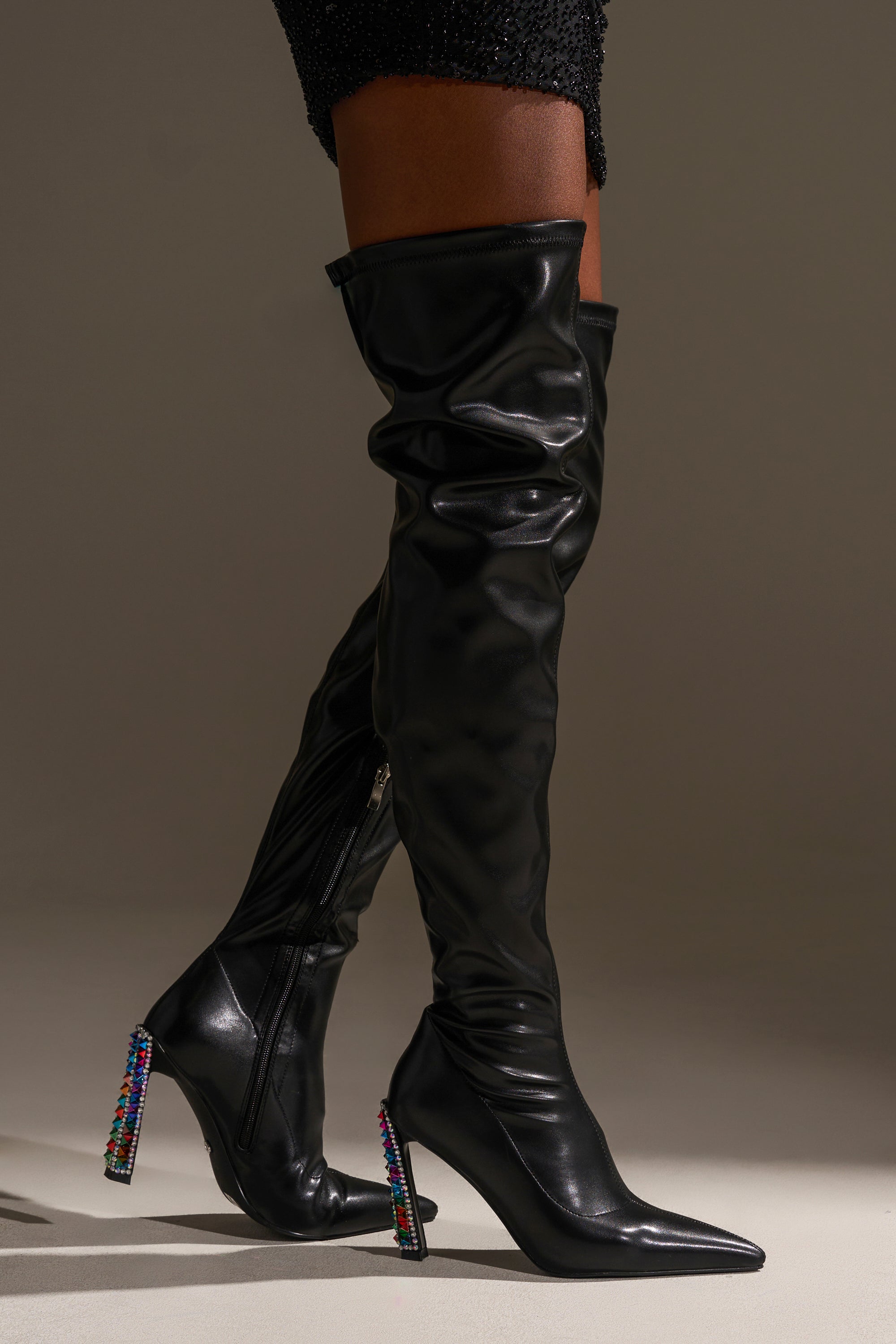 A person wearing the AZALEA WANG COSMIC BLING BLACK BOOT WITH EMBELLISHED HEEL—slouchy, over-the-knee boots with pointed toes and jeweled block heels—paired with a black textured mini skirt.