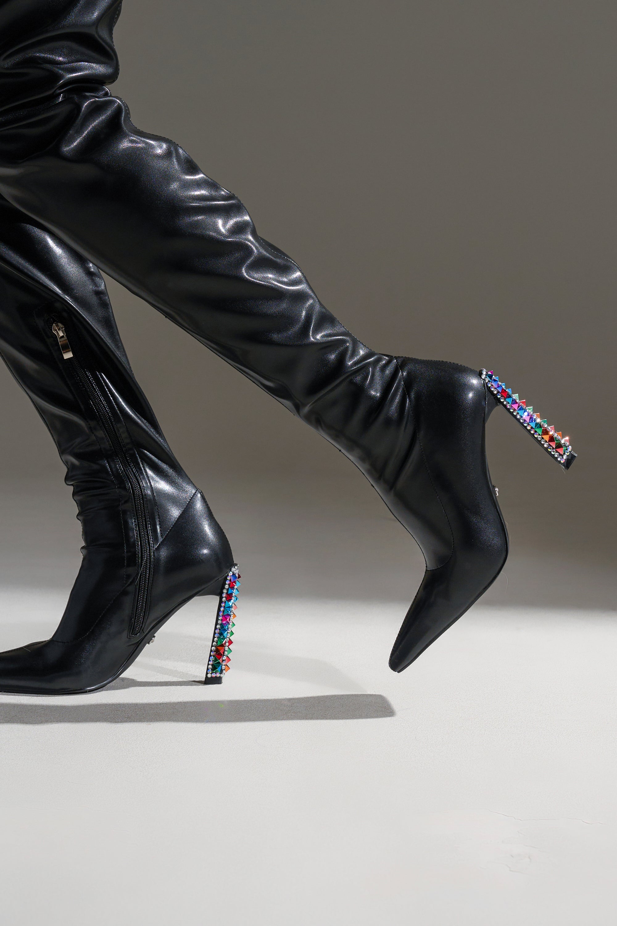 A person models the AZALEA WANG COSMIC BLING BLACK BOOT WITH EMBELLISHED HEEL—black, pointed-toe, faux leather knee-high boots with silver zippers and gem-decorated block heels—striding against a neutral backdrop.