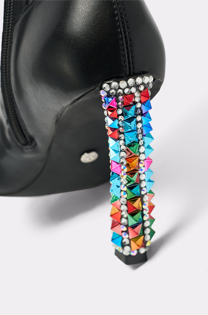 The AZALEA WANG COSMIC BLING BLACK BOOT features black faux leather, a stiletto heel embellished with colorful metallic studs and silver rhinestones, shown against a plain light background.