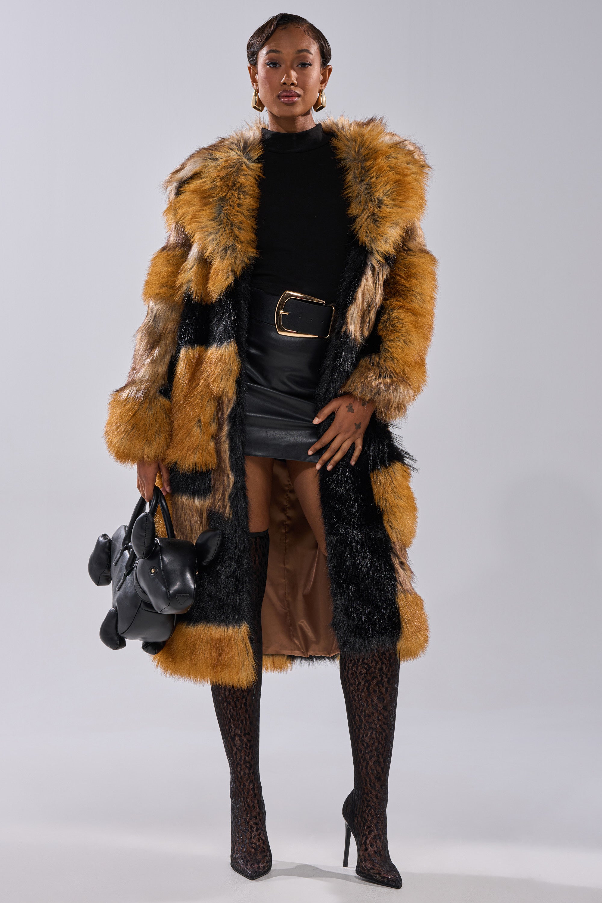 A stylish woman wears the COSMO PATCHWORK FAUX FUR coat with orange and black stripes over a black turtleneck and leather mini skirt, paired with patterned tights, high heels, and a black handbag.