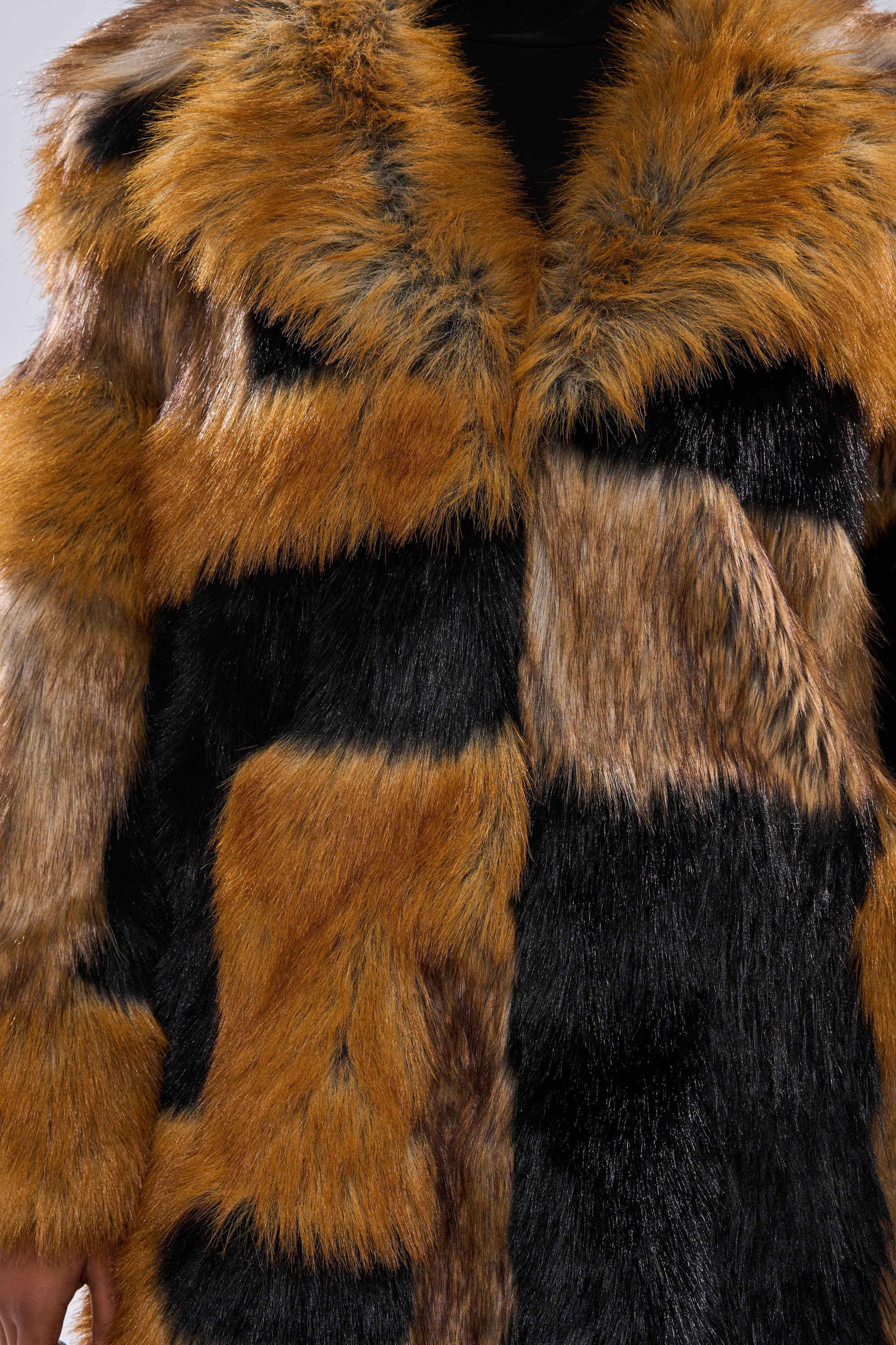 A close-up shows a person wearing the COSMO PATCHWORK FAUX FUR, a multi-colored jacket with brown, black, and tan faux fur patches in a fluffy, textured finish. Only the torso and part of one arm are visible.