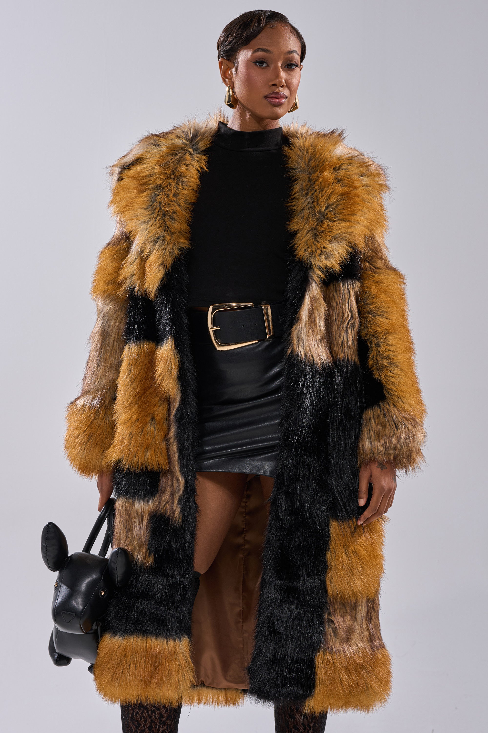 A woman wears the COSMO PATCHWORK FAUX FUR coat in brown, black, and gold over a black turtleneck and mini skirt. She accessorizes with a black animal-head bag, styled short hair, and gold hoop earrings.