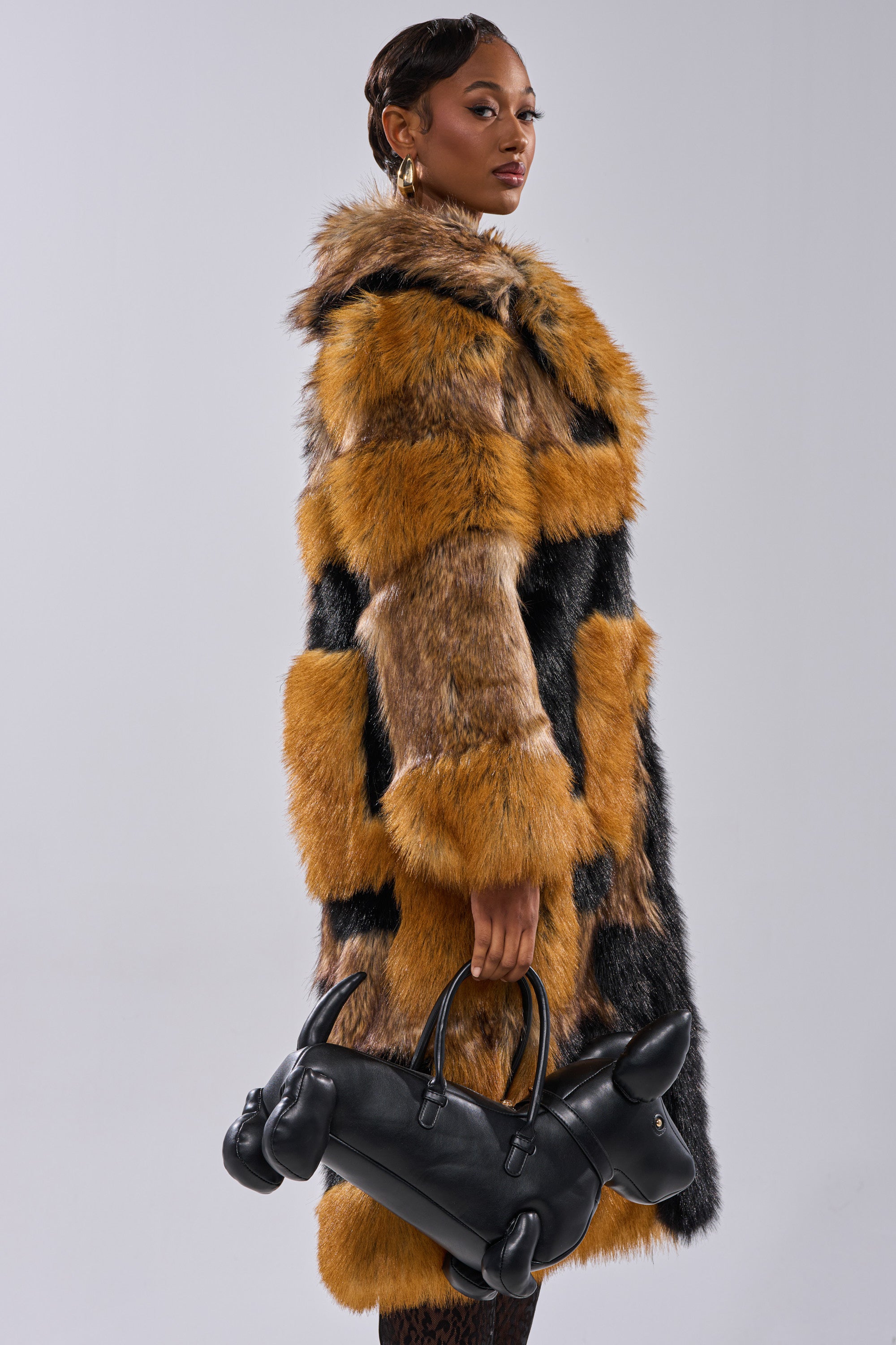 A woman wears the COSMO PATCHWORK FAUX FUR jacket in brown and black, standing against a plain background while holding a black dog-shaped handbag.