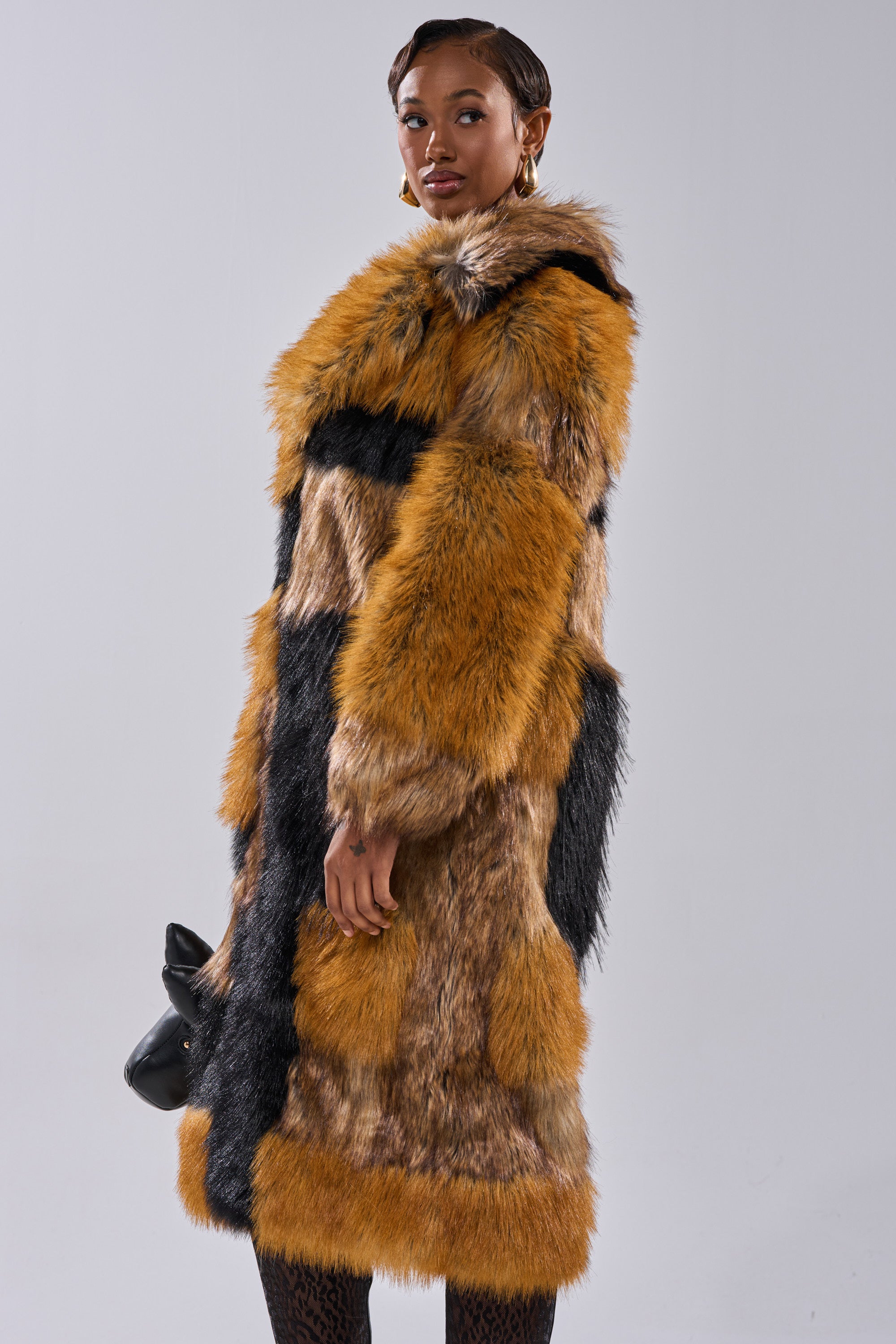 A woman models the COSMO PATCHWORK FAUX FUR, a long, luxurious jacket in brown, black, and tan patches. She stands against a gray background holding a black handbag, gazing confidently at the camera.
