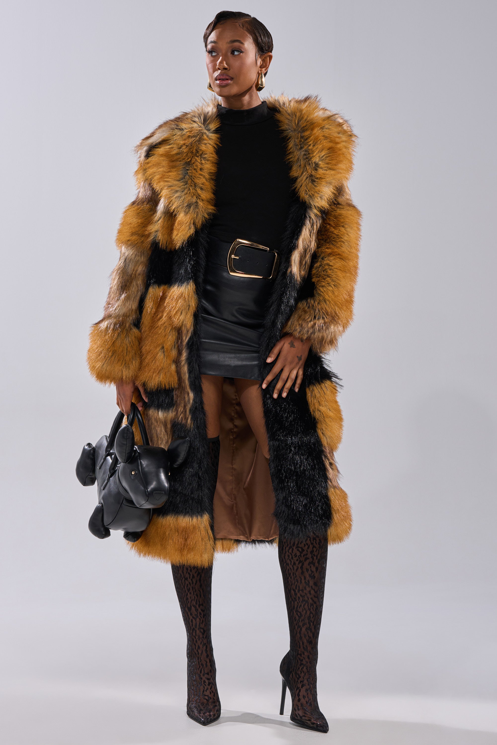 A stylish person wears the COSMO PATCHWORK FAUX FUR coat with black and orange stripes, a black top, belted leather mini skirt, patterned thigh-high boots, and a black handbag, standing confidently against a plain background.