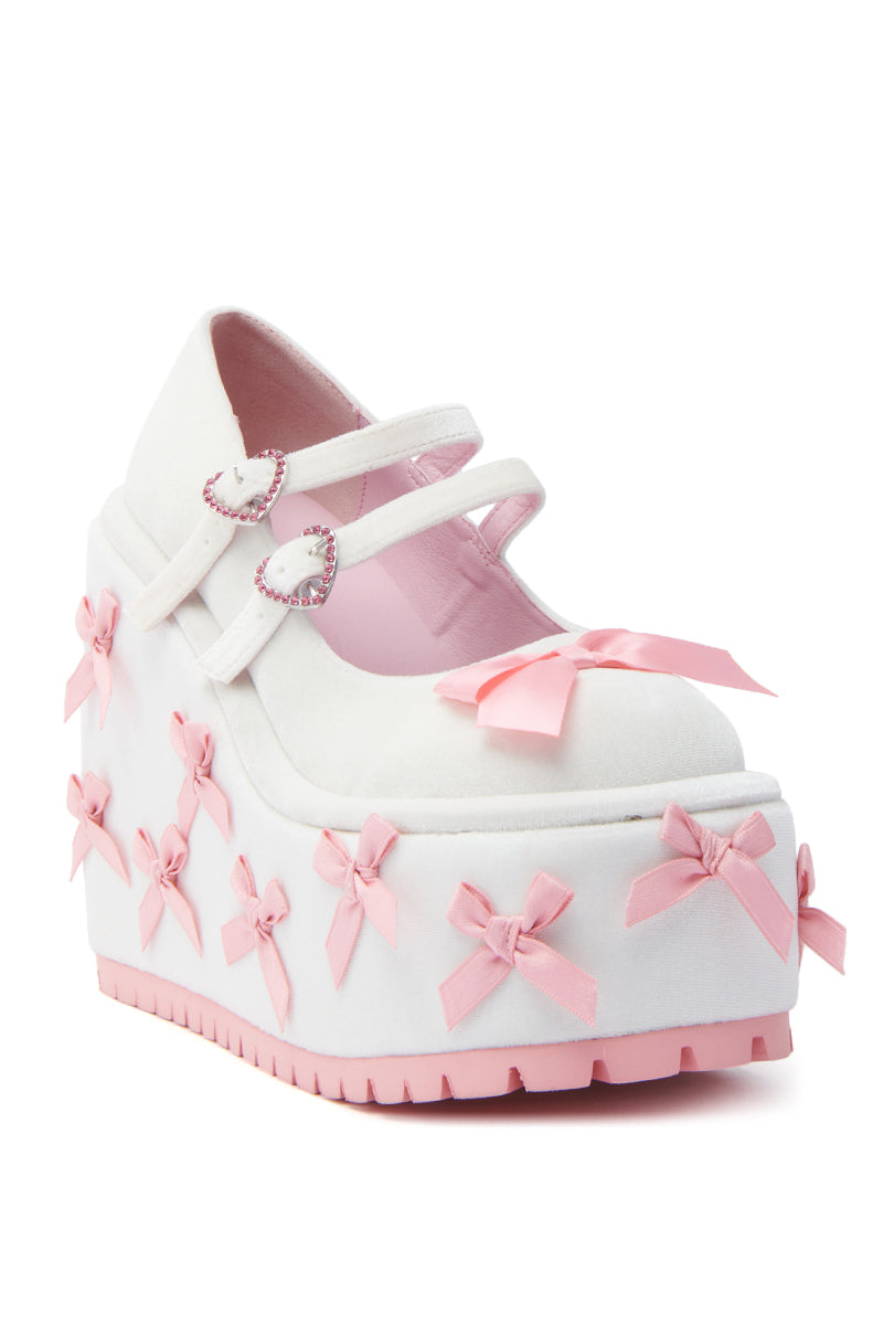 The KOI FOOTWEAR A COURTSHIP OF BOWS PINK WEDGE SNEAKER features a velvet upper, thick pink platform wedge, multiple pink bows, and two heart-shaped buckle straps for a playful and elegant look.