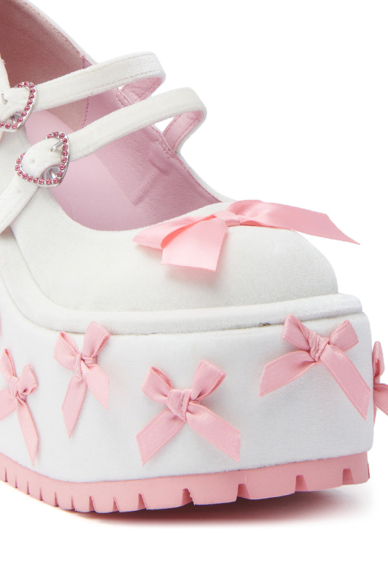A close-up of the KOI FOOTWEAR A COURTSHIP OF BOWS PINK WEDGE SNEAKER, featuring Mary Jane straps with heart-shaped buckles, pink bows, a pink platform wedge sole with ribbon accents, a velvet upper, and soft pink lining.