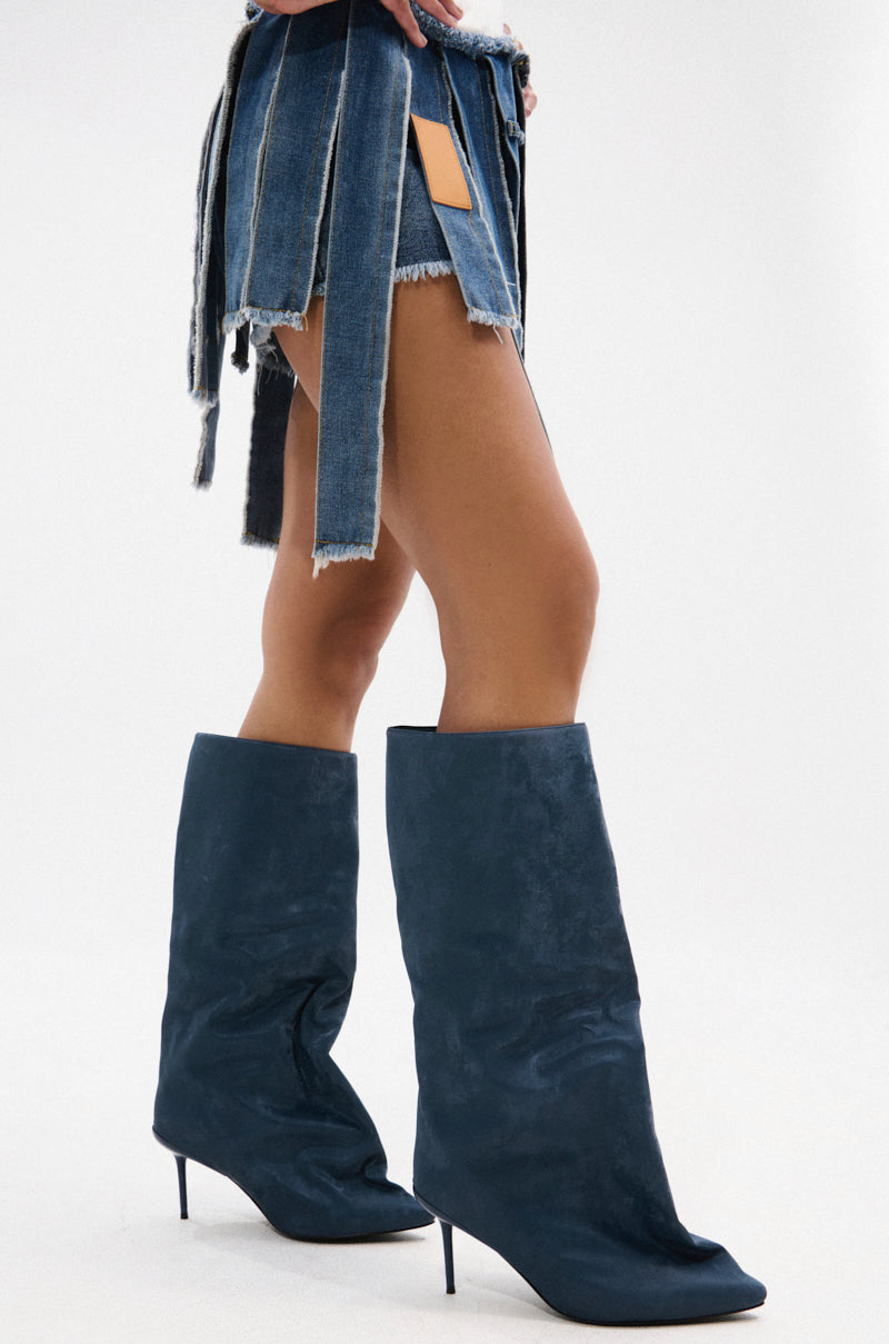 A person models the AZALEA WANG COUTURE CRAZY BLUE LUXE SHINE LOOK BOOT—tall, slouchy navy stiletto heels—paired with a short, distressed denim skirt with frayed edges, standing against a plain white background.