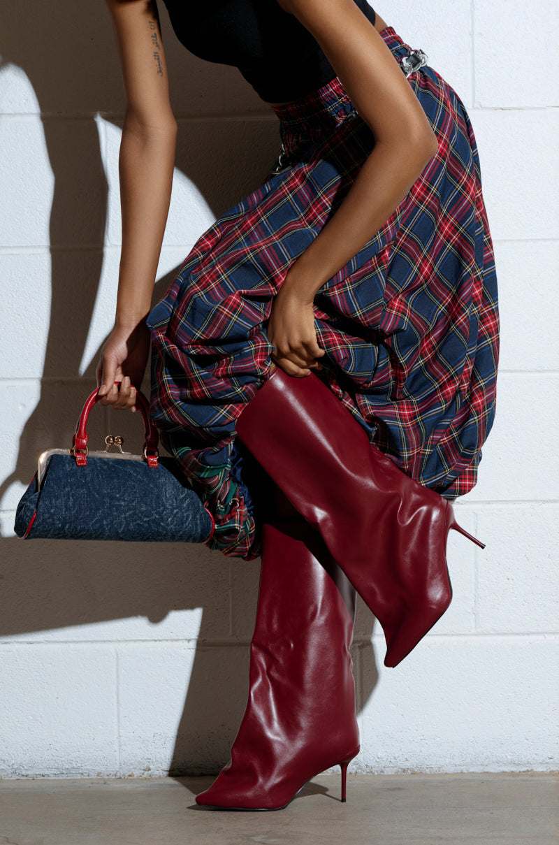 Wearing a plaid skirt and AZALEA WANG COUTURECRAZY BURGUNDY BOOTS, a person bends down, holding a small dark blue handbag against a white textured wall, enhancing the vibrant outfit colors.