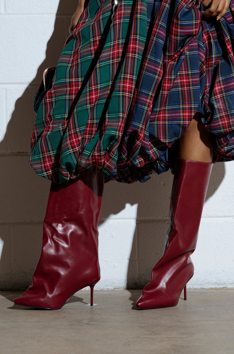 Against a white brick wall, a person wears a voluminous plaid skirt in blue, red, and green and knee-high AZALEA WANG COUTURECRAZY burgundy boots with pointed toes and stiletto heels.