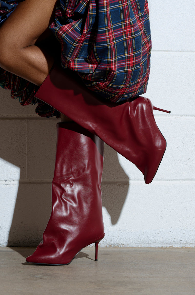 Seated against a white wall, a person showcases the AZALEA WANG COUTURECRAZY BURGUNDY BOOT—a striking knee-high, pointed-toe stiletto in faux red leather—perfectly paired with a blue, burgundy, and yellow plaid skirt, highlighting both the intricate boot and skirt details.