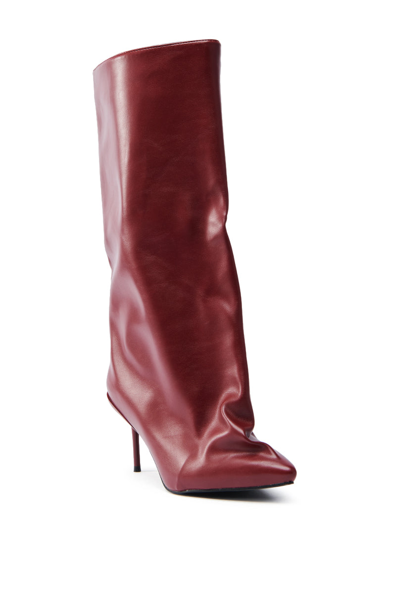 The AZALEA WANG COUTURECRAZY BURGUNDY BOOT flaunts a stiletto heel, slouchy shaft, and pointed toe. Made of glossy faux leather, it stands against a white background highlighting its elegant design.
