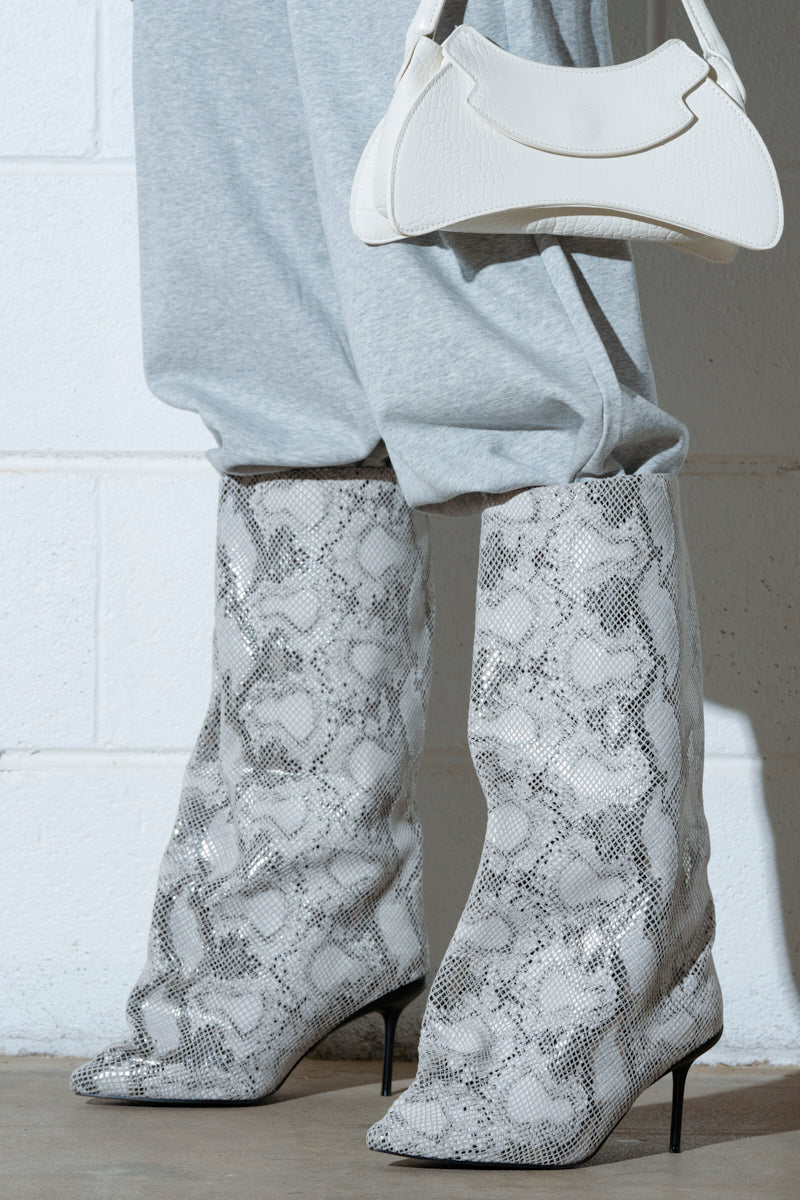 Someone wears the AZALEA WANG COUTURECRAZY SNAKE BOOT knee-highs with gray sweatpants, elegantly holding a small white handbag. They pose confidently against a white brick wall, showcasing their unique style.