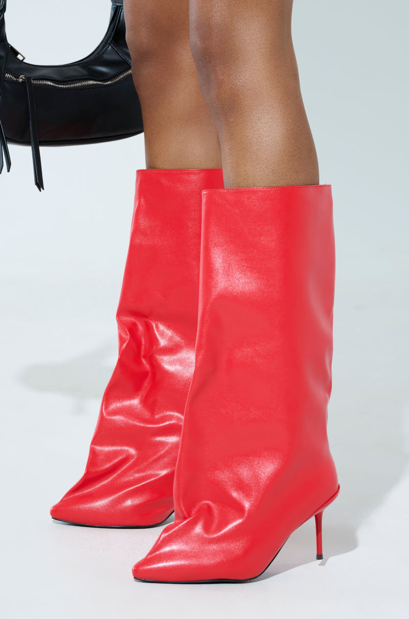 A person wearing the AZALEA WANG COUTURE CRAZY RED BOOT, shiny and oversized with mid-calf length, pointed toes, and stiletto heels, is holding a small black handbag against a plain background.