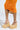 The image shows a person in a bright orange ruched dress and AZALEA WANG COVA BEIGE WEDGE sandals. The focus is on the lower half of the outfit against a plain background, highlighting the elegant summer shoes with their stylish platform sole.