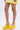 A person stands against a plain white background, showcasing their legs and the AZALEA WANG COVA YELLOW WEDGE shoes styled with a short yellow skirt. The focus is on the footwear.