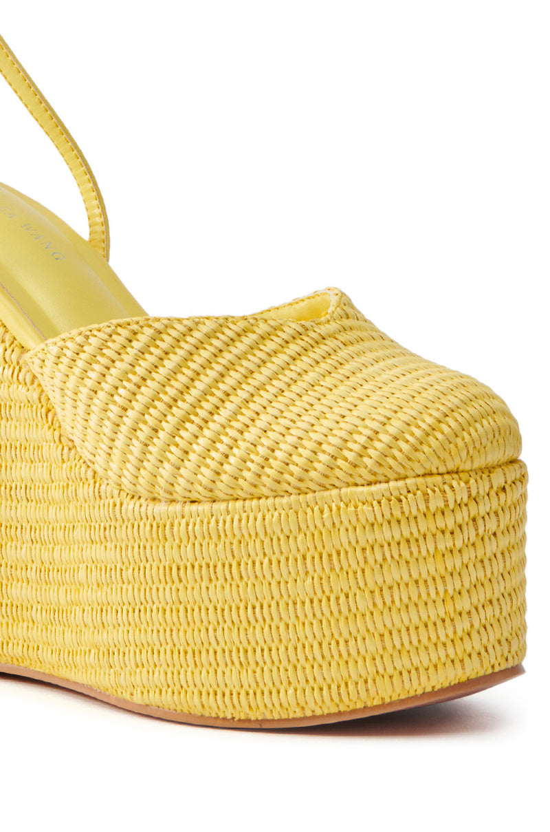 Close-up of the AZALEA WANG COVA YELLOW WEDGE, featuring a thick platform sole and closed toe. The wicker-inspired texture has a straw-like feel. A strap from the heel suggests a slingback style, with a flat sole and subtle heel elevation.