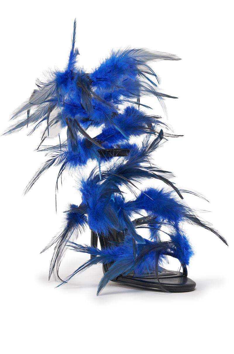 The AKIRA GOLD COVERT FEATHER EMBELLISHED SPIRAL STRAP STILETTO SANDAL features striking blue and black feathers for a bold, avant-garde look, dramatically set against a plain white background. 5
