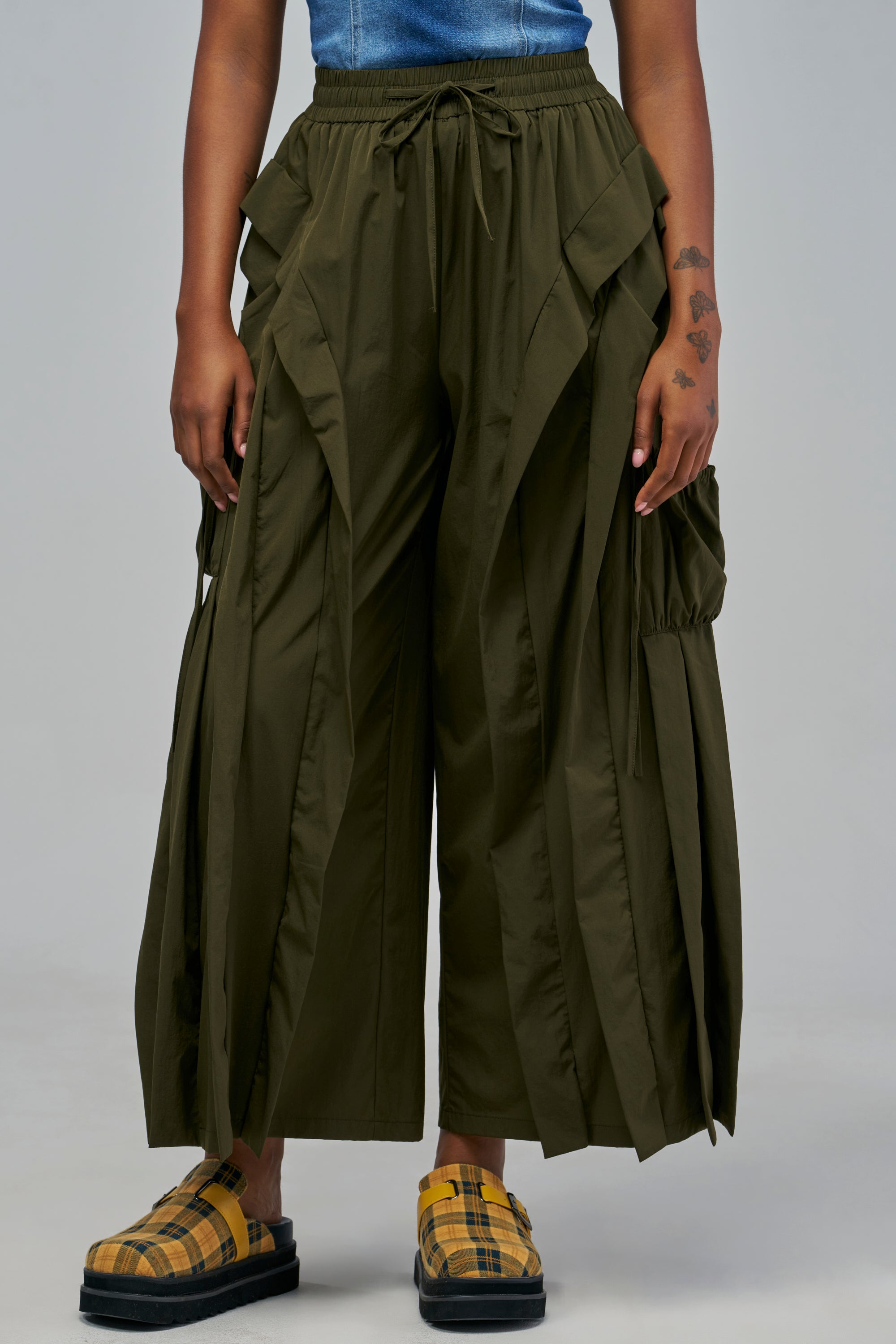 GREEN ENVY PANT olive green wide-leg cargo pants with a drawstring waist and layered pleated panels, styled with plaid slide sandals for a streetwear look.