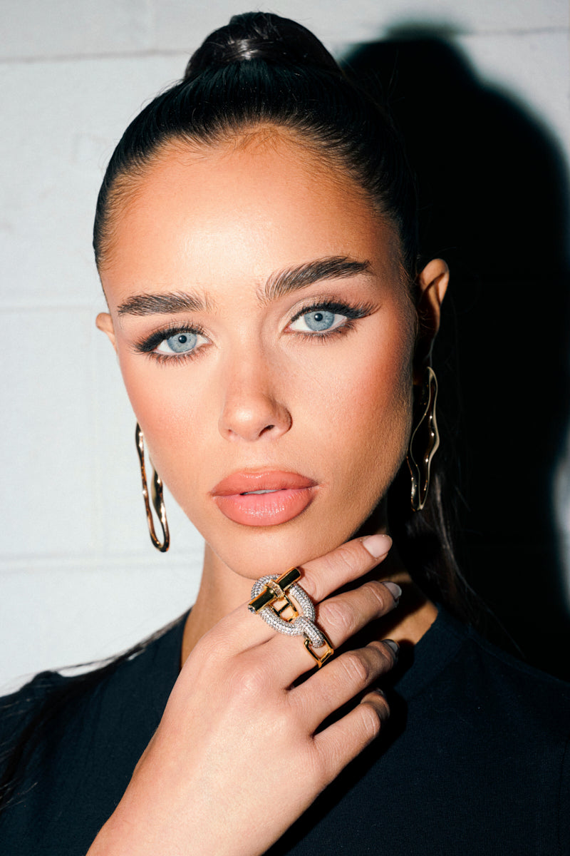 A woman with blue eyes and sleek dark hair in a ponytail poses against a light background, wearing gold earrings and the LUXE LIFE COCKTAIL RING—her hand gently touching her chin and showcasing its sparkling rhinestone embellishments.