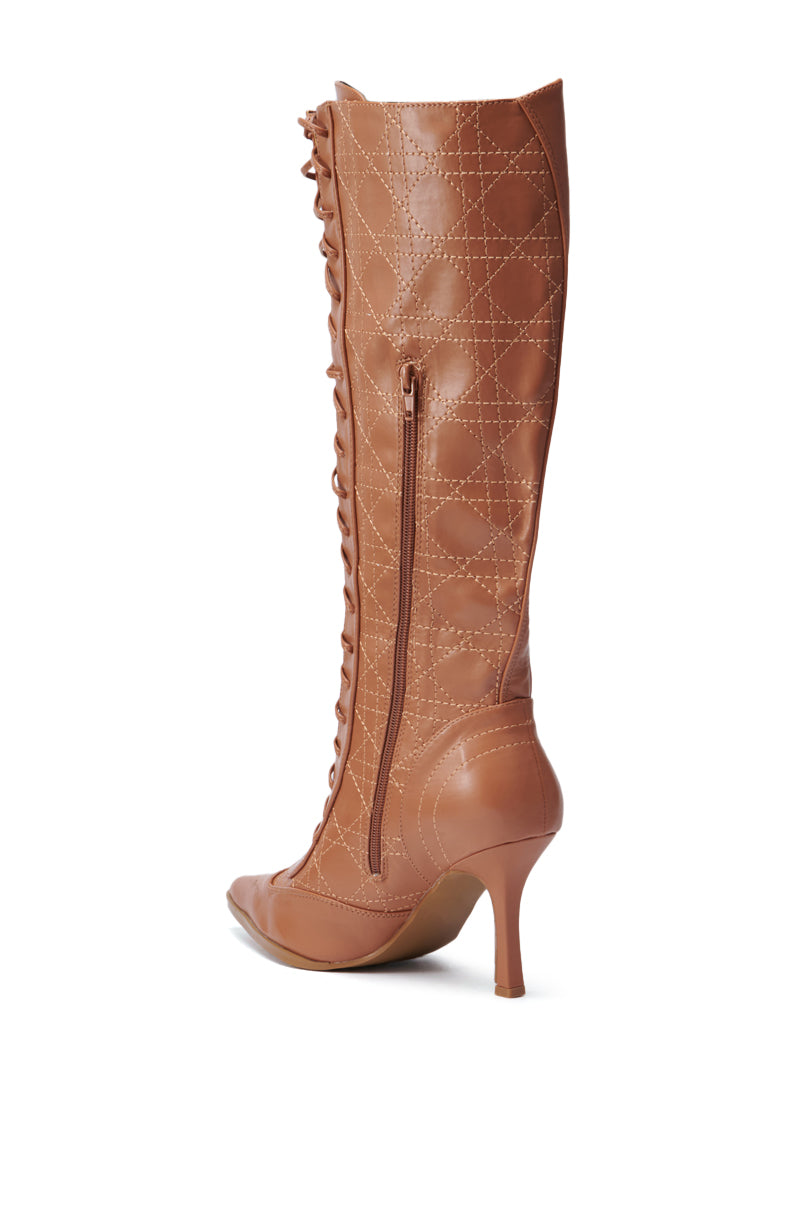 The CREATOR TAN LACE UP STILETTO BOOT is displayed against a white background, highlighting its knee-high shaft in a beige faux leather design. It features intricate quilted stitching, a side zipper, and an elegant lace-up detail at the front.