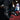 A person wearing black faux leather pants with frayed hems and AZALEA WANG CRESSINDA BLACK GUNMETAL DOME STUD STILETTO BOOTIES rests their legs on a red upholstered chair in a dimly lit room.