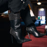 A person wearing black faux leather pants with frayed hems and AZALEA WANG CRESSINDA BLACK GUNMETAL DOME STUD STILETTO BOOTIES rests their legs on a red upholstered chair in a dimly lit room.
