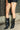 A person wearing black pinstriped shorts and AZALEA WANG CRESSINDA BLACK GUNMETAL DOME STUD STILETTO BOOTIES stands on a sidewalk by a brick wall and metal fence, with one hand resting by their side.
