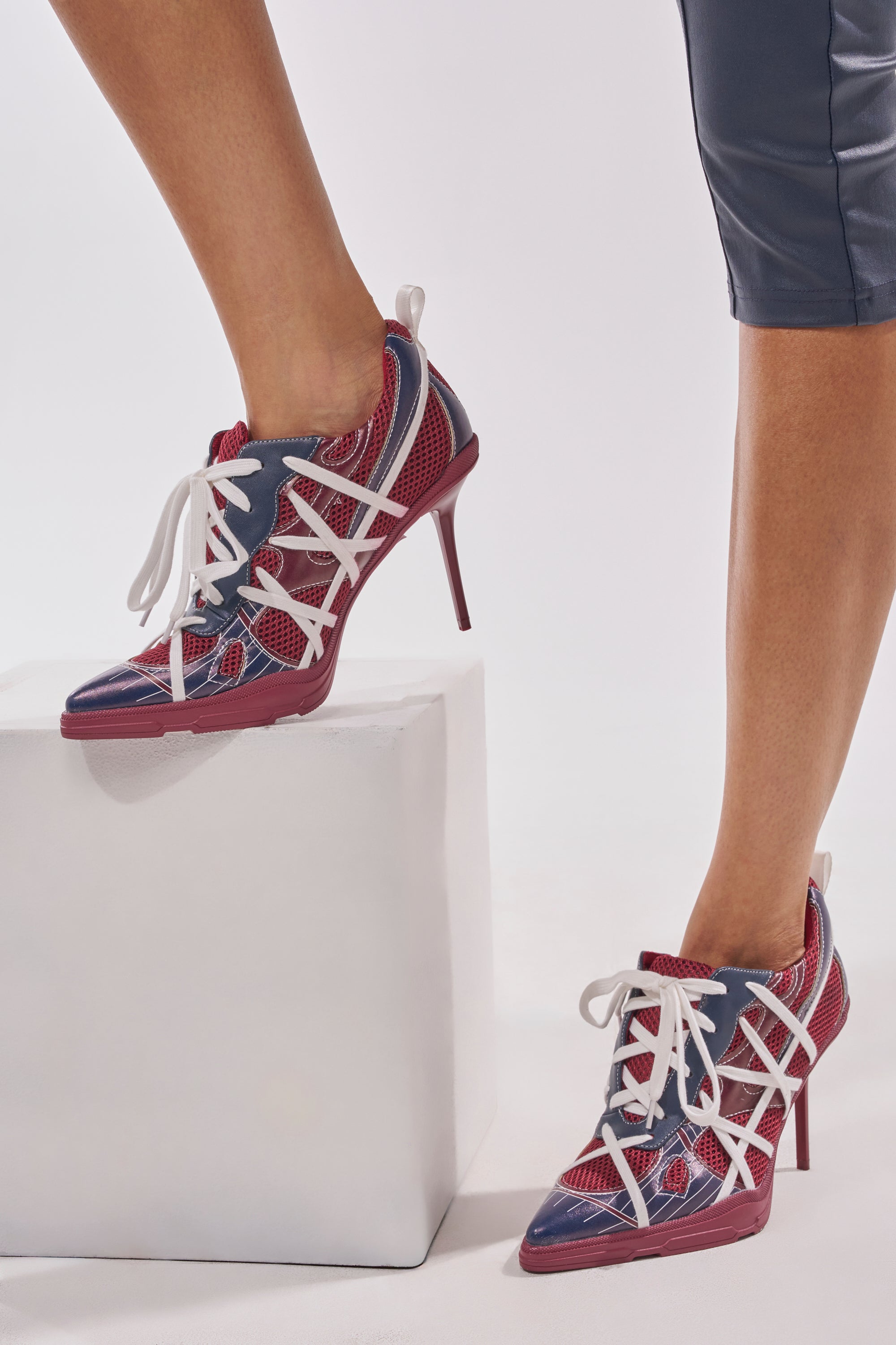 A person in dark fitted pants models the AZALEA WANG CROSS WALK BURGUNDY STILETTO SNEAKER HEEL, featuring sporty-chic geometric cutouts, with one foot on a white block and the other on the floor. 0