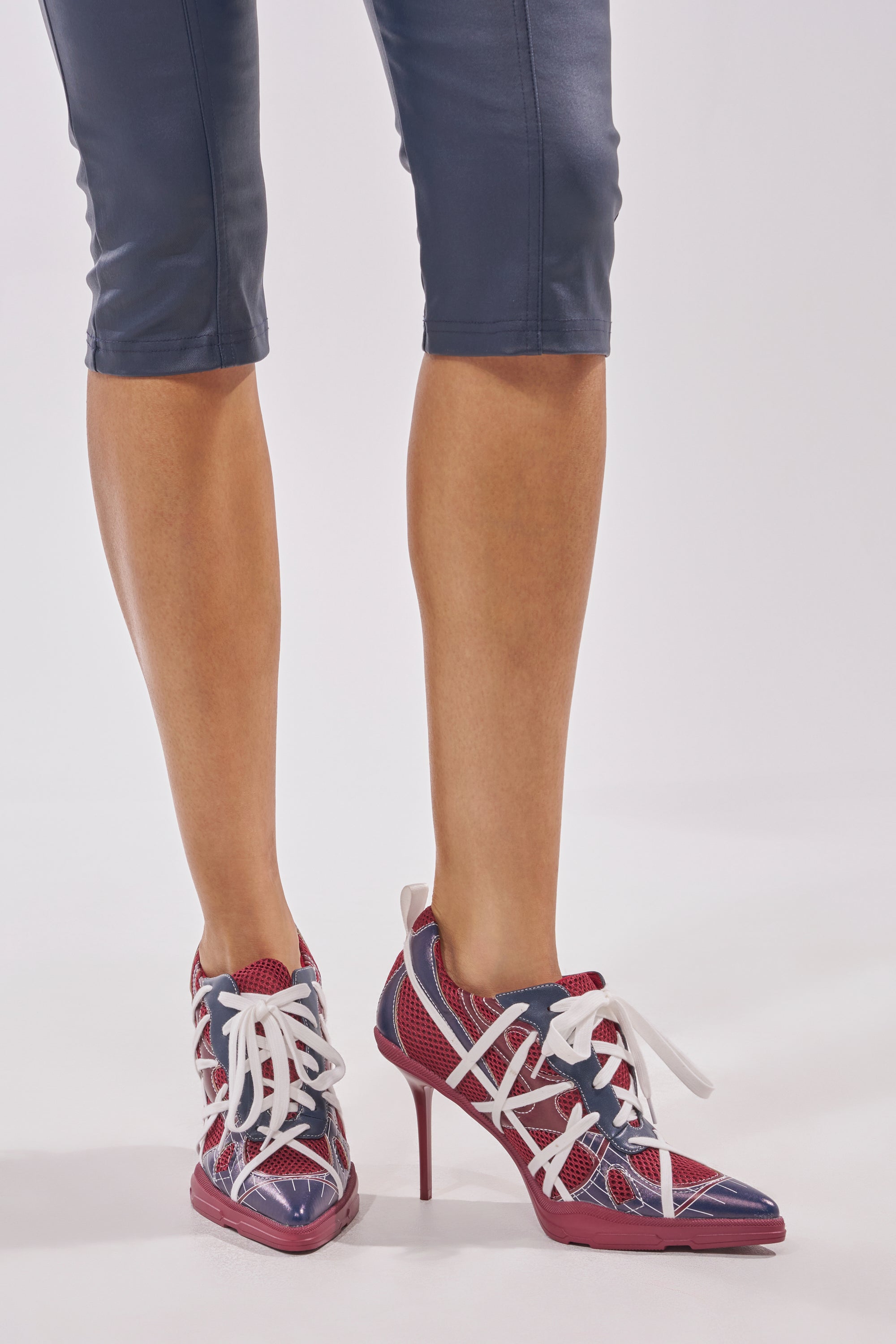 A person wears blue cropped pants and the AZALEA WANG CROSS WALK BURGUNDY STILETTO SNEAKER HEEL—sporty-chic lace-up stiletto heels styled like sneakers—while standing on a white background. 1
