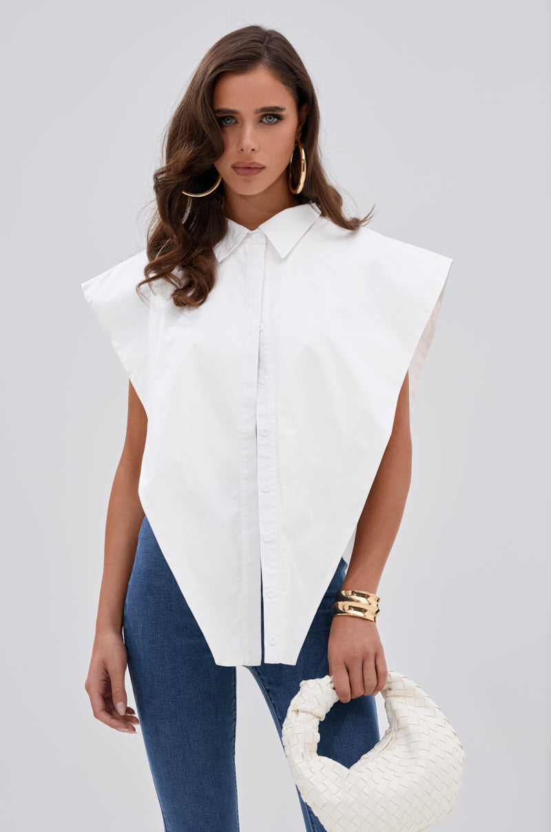 A woman with long brown hair wears the ALWAYS GIVING BUTTON DOWN POPLIN BLOUSE, blue jeans, hoop earrings, gold bracelets, and holds a white woven handbag. She stands against a plain light gray background. 0