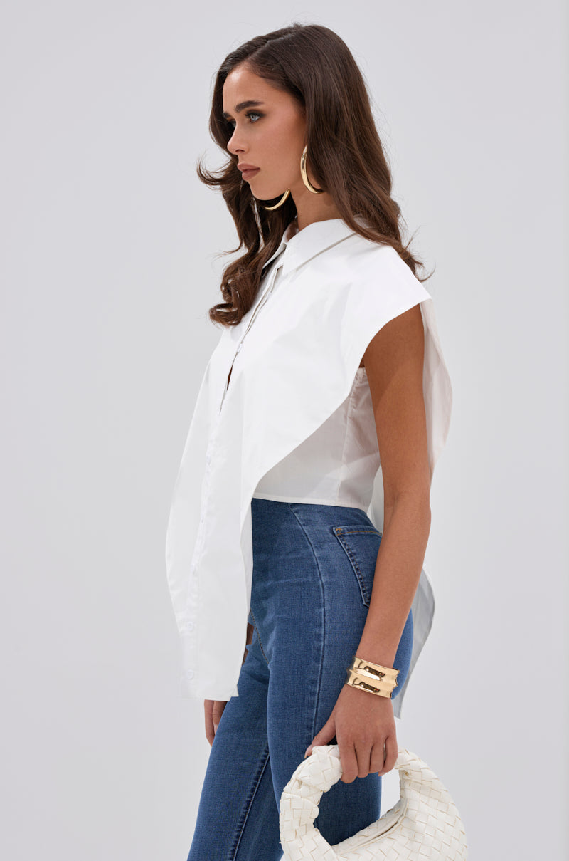 A woman with long brown hair wears the ALWAYS GIVING BUTTON DOWN POPLIN BLOUSE, styled sleeveless and asymmetrical in white, paired with blue jeans, gold hoop earrings and bracelets, holding a white woven handbag against a plain light background. 1