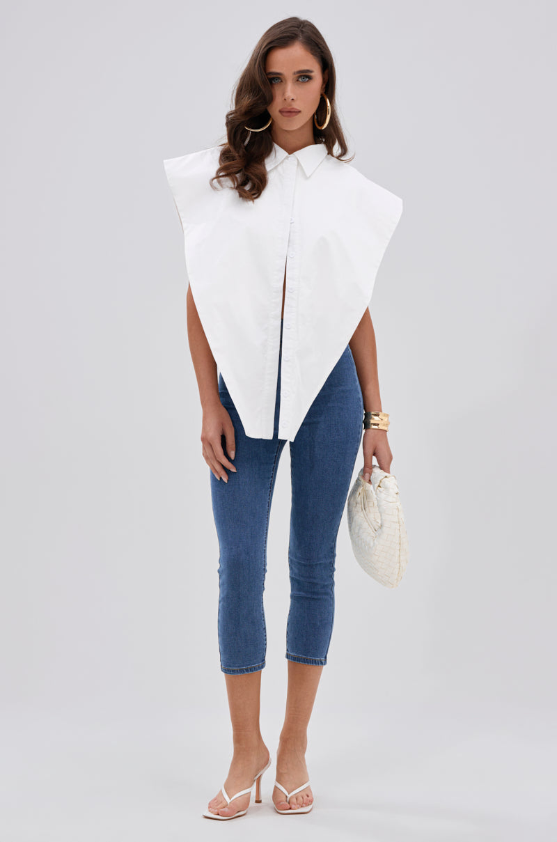 A woman stands against a plain background wearing the ALWAYS GIVING BUTTON DOWN POPLIN BLOUSE—an oversized white sleeveless collared shirt—paired with blue cropped skinny jeans, white heeled sandals, a textured white handbag, gold hoop earrings, and bracelets. 3