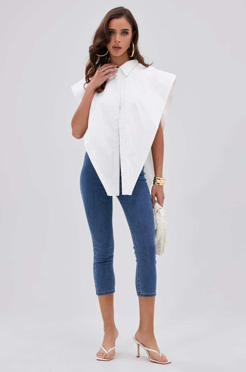 A woman wearing the ALWAYS GIVING BUTTON DOWN POPLIN BLOUSE, blue cropped skinny jeans, white heeled sandals, gold jewelry, and holding a white textured clutch stands against a plain light gray background. 4
