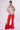 A person wearing the SHINE TOO BRIGHT TEE with an oversized boxy silhouette, vibrant red pants with white side stripes, and large, fluffy red slippers stands against a plain background. They are accessorized with hoop earrings and clear glasses.