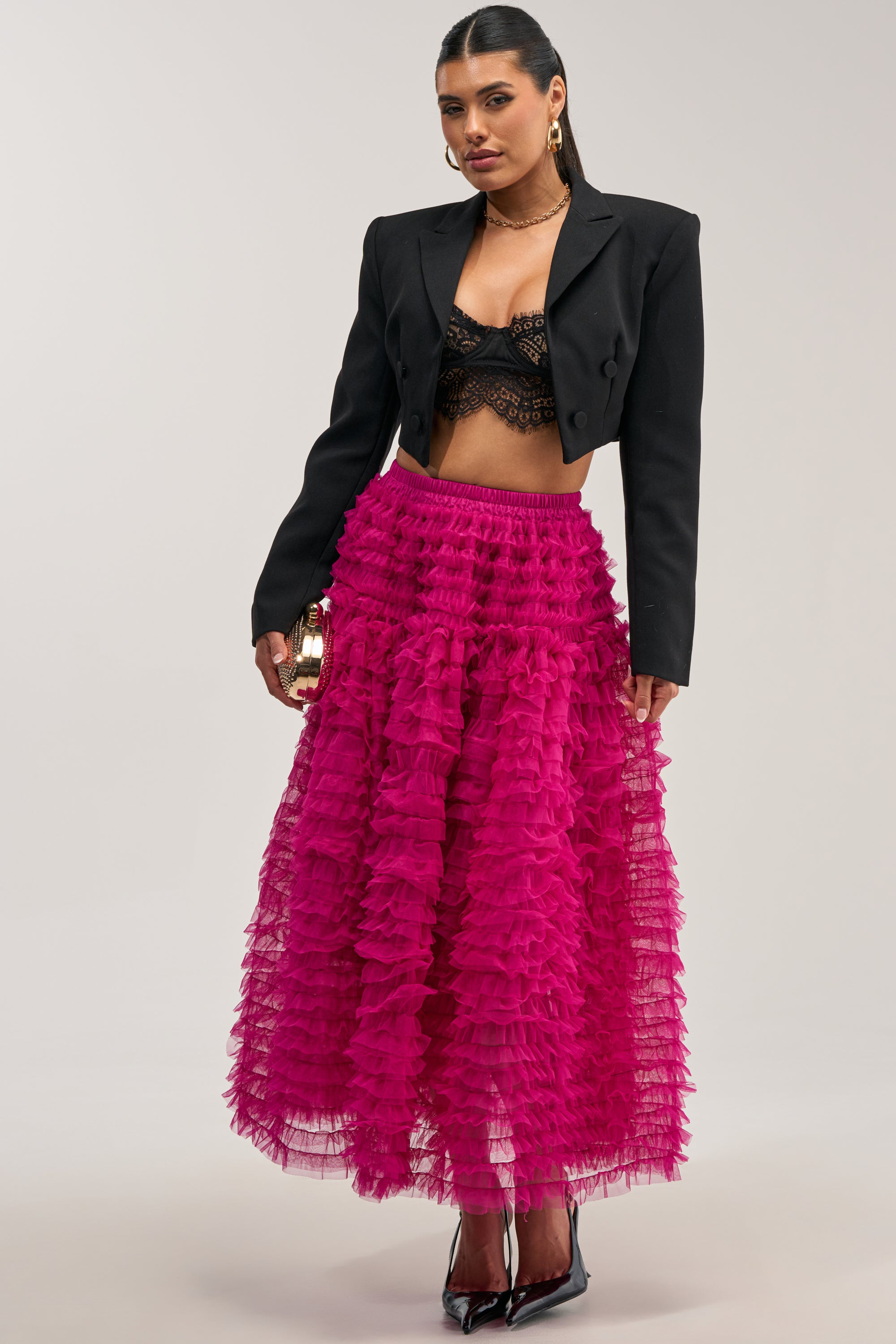 A woman wears a black cropped blazer, black lace bralette, black heels, gold hoop earrings, and holds a small gold clutch. Her hair is slicked back and she strikes a confident pose—the LOVE THE DRAMA MESH MAXI SKIRT IN FUCHSIA ensures all eyes are on her for date night.