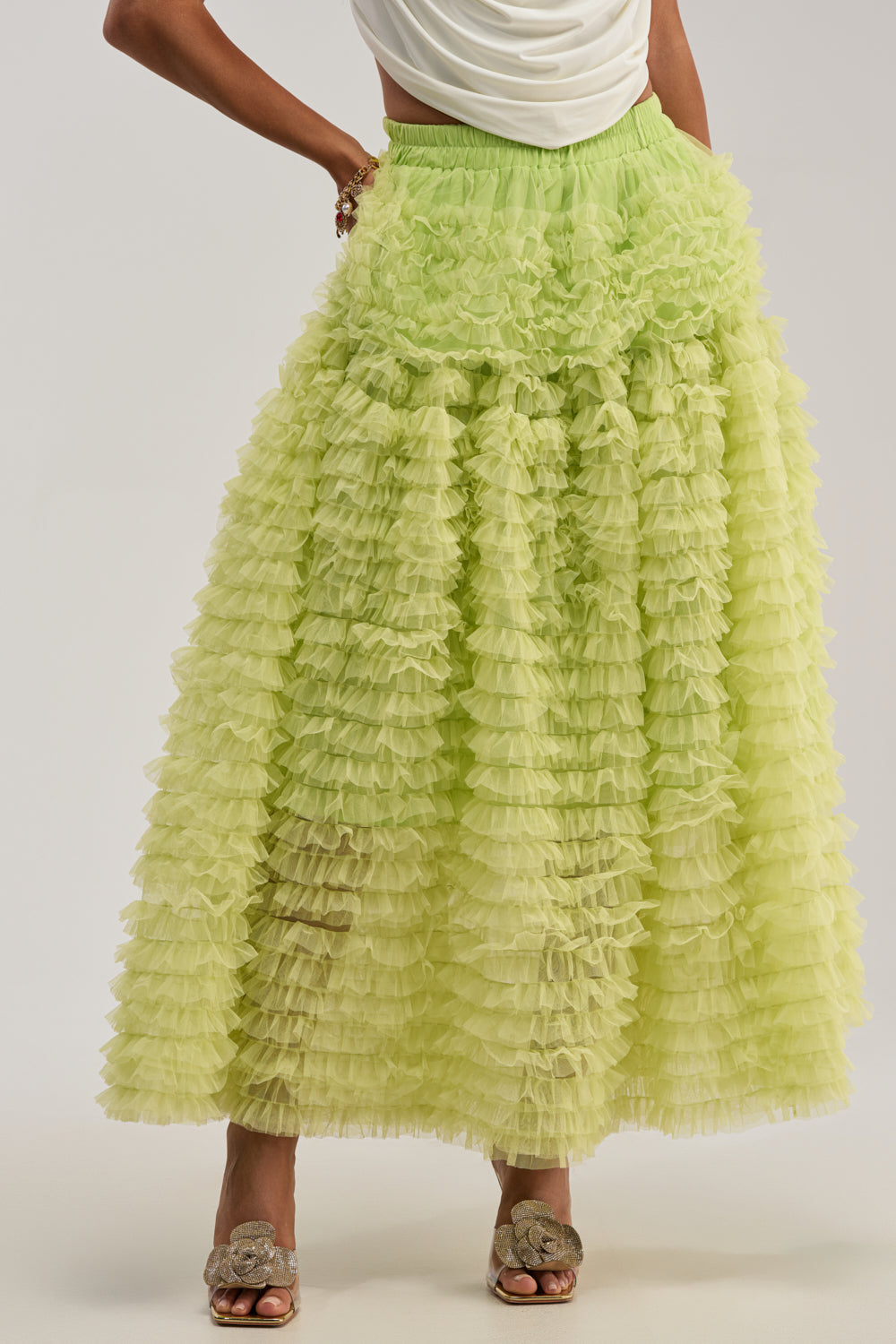 Wearing the LOVE THE DRAMA MESH MAXI SKIRT in lime green with silver open-toe shoes, a person poses with one hand on their hip. Their white top is only partially visible.