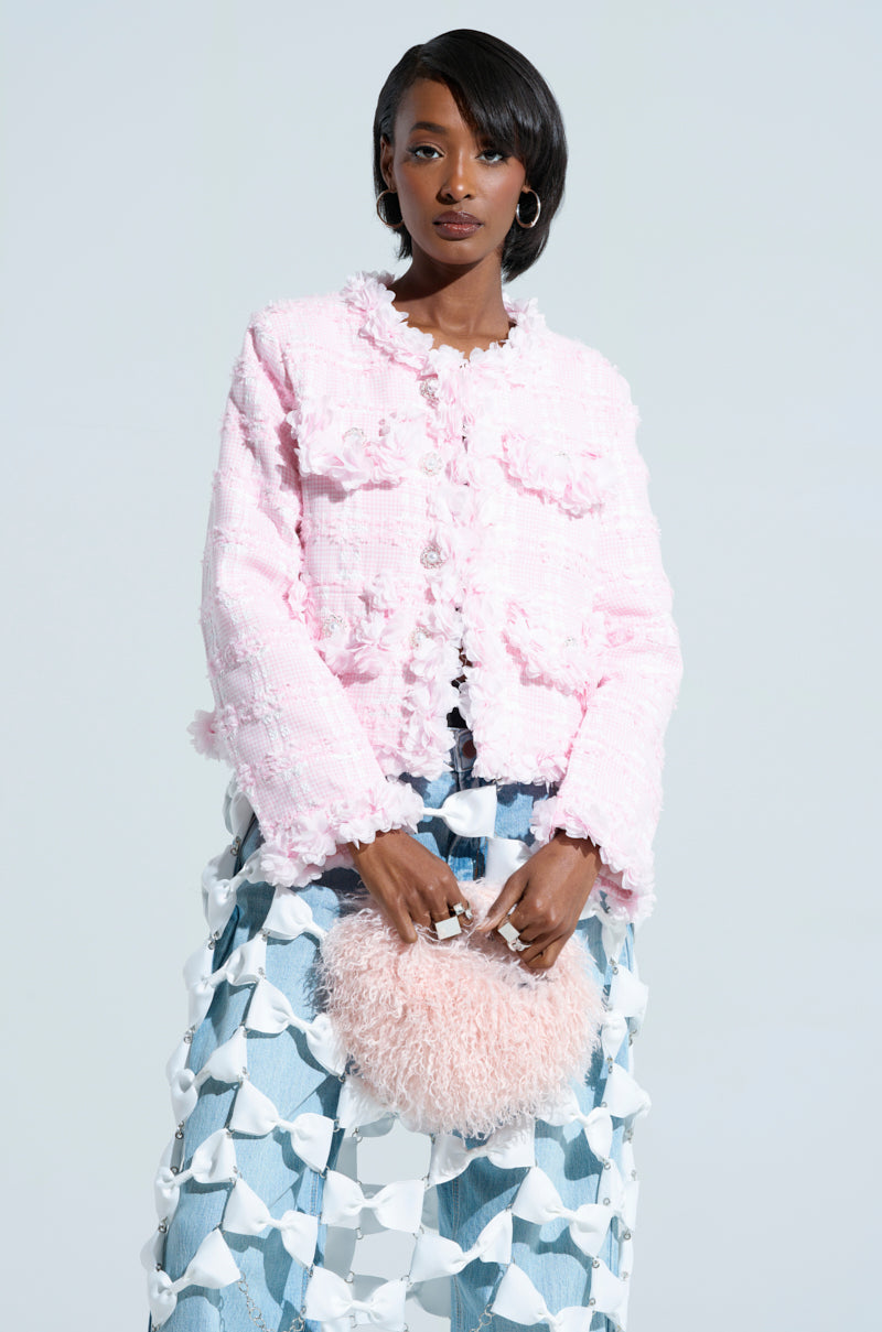 A woman stands against a light background, wearing the SPRINGTIME TWEED BLAZER with floral appliques and a blue patterned skirt. She holds a fluffy pink handbag and has short, styled hair.