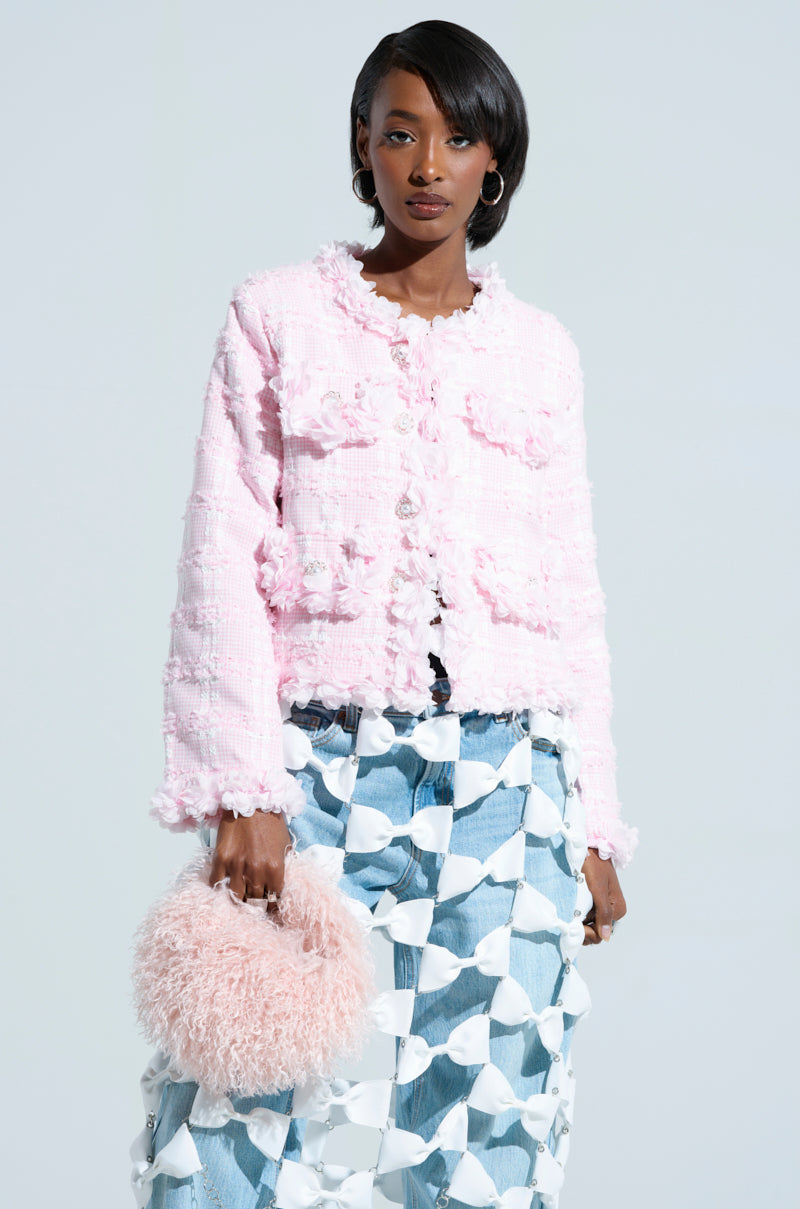 A person wearing the SPRINGTIME TWEED BLAZER paired with light blue patterned pants holds a fluffy pink handbag. The plain light gray background adds subtle contrast to this springtime ensemble.