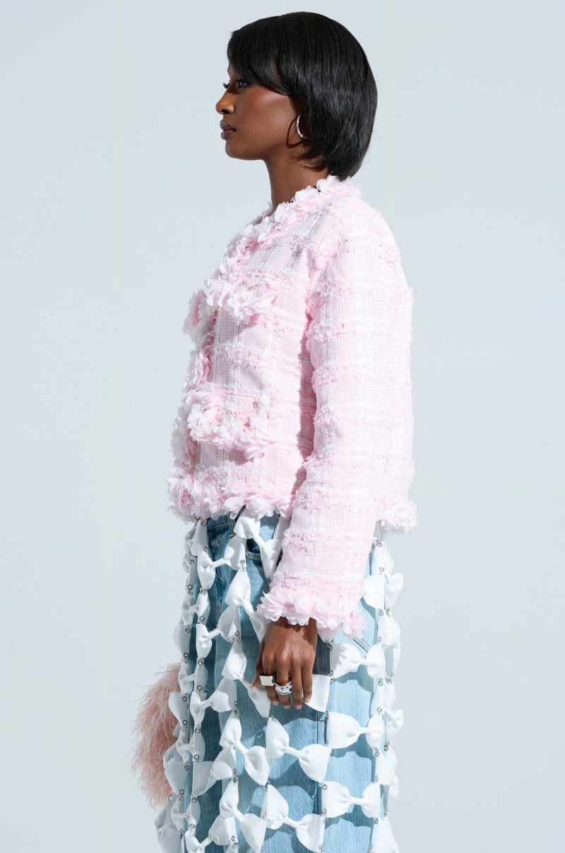 A person with short black hair stands in profile against a light background. They are wearing a SPRINGTIME TWEED BLAZER and a blue skirt decorated with floral appliques, holding a fluffy pink object.