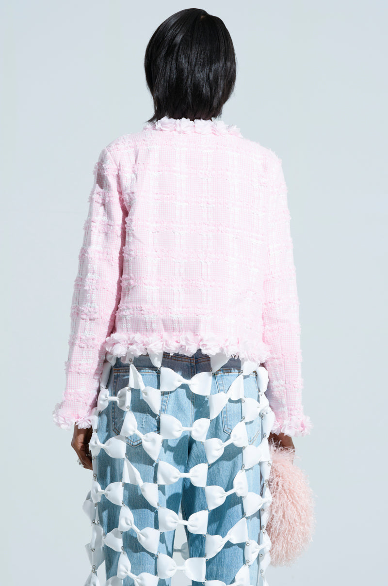 Rear view of a person wearing the SPRINGTIME TWEED BLAZER, characterized by its textured design, paired with a creative skirt made of white interlocking loops over faded denim jeans. They hold a pink, fluffy accessory in their hand against a plain background.