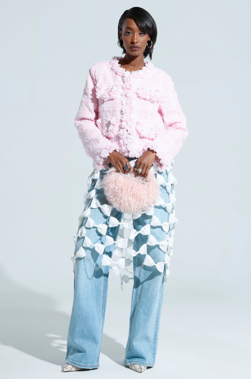 A stylish individual poses against a plain backdrop, wearing the SPRINGTIME TWEED BLAZER in textured pink, paired with an eye-catching geometric skirt layered over wide-leg jeans. They carry a fluffy pink purse embellished with floral appliques and complete the look with white high-heeled shoes.