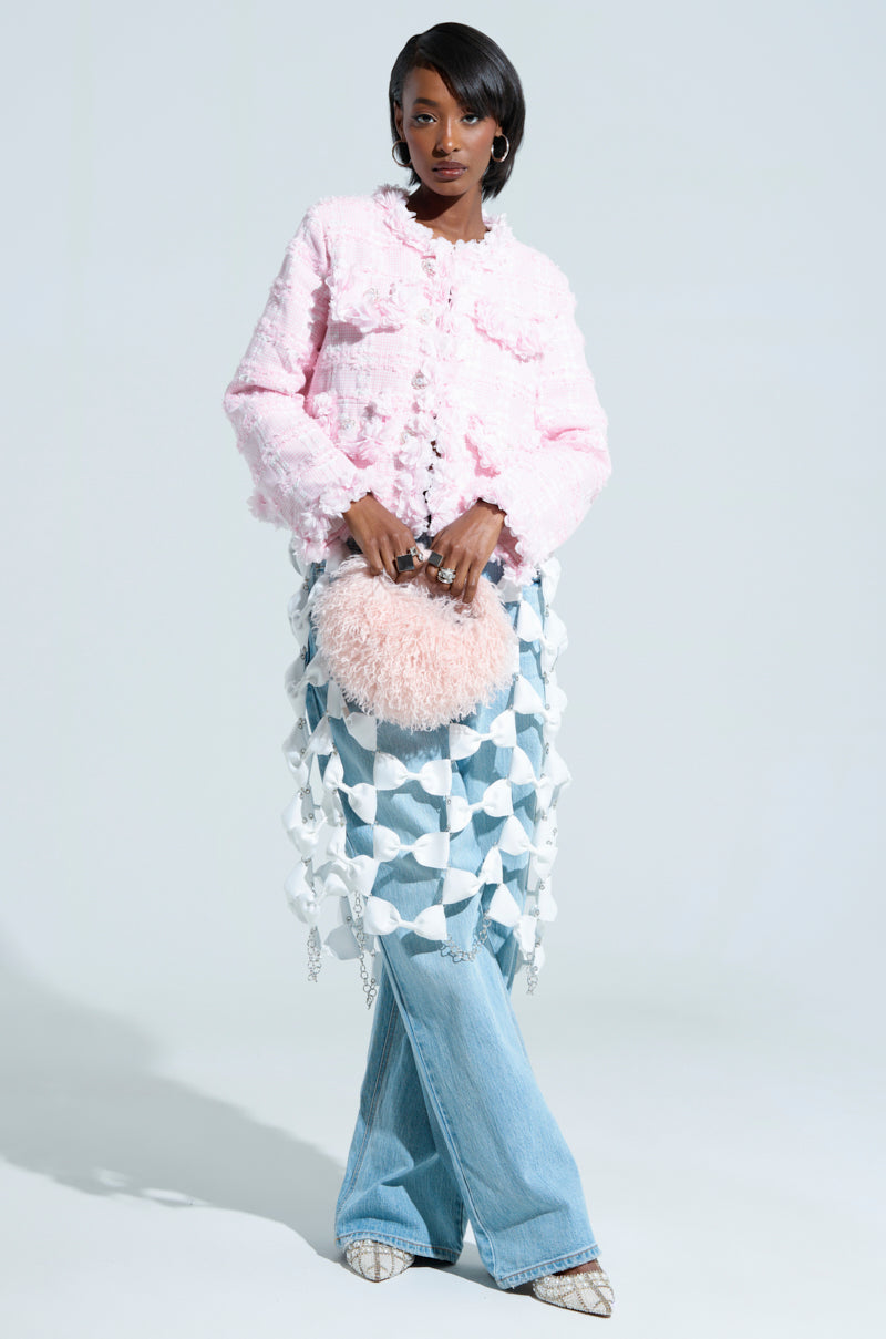 A person poses in a stylish outfit showcasing a light pink SPRINGTIME TWEED BLAZER, paired with a geometric skirt overlay on blue jeans. They hold a fluffy pink bag adorned with floral appliques and wear patterned shoes, set against a plain light-colored background.
