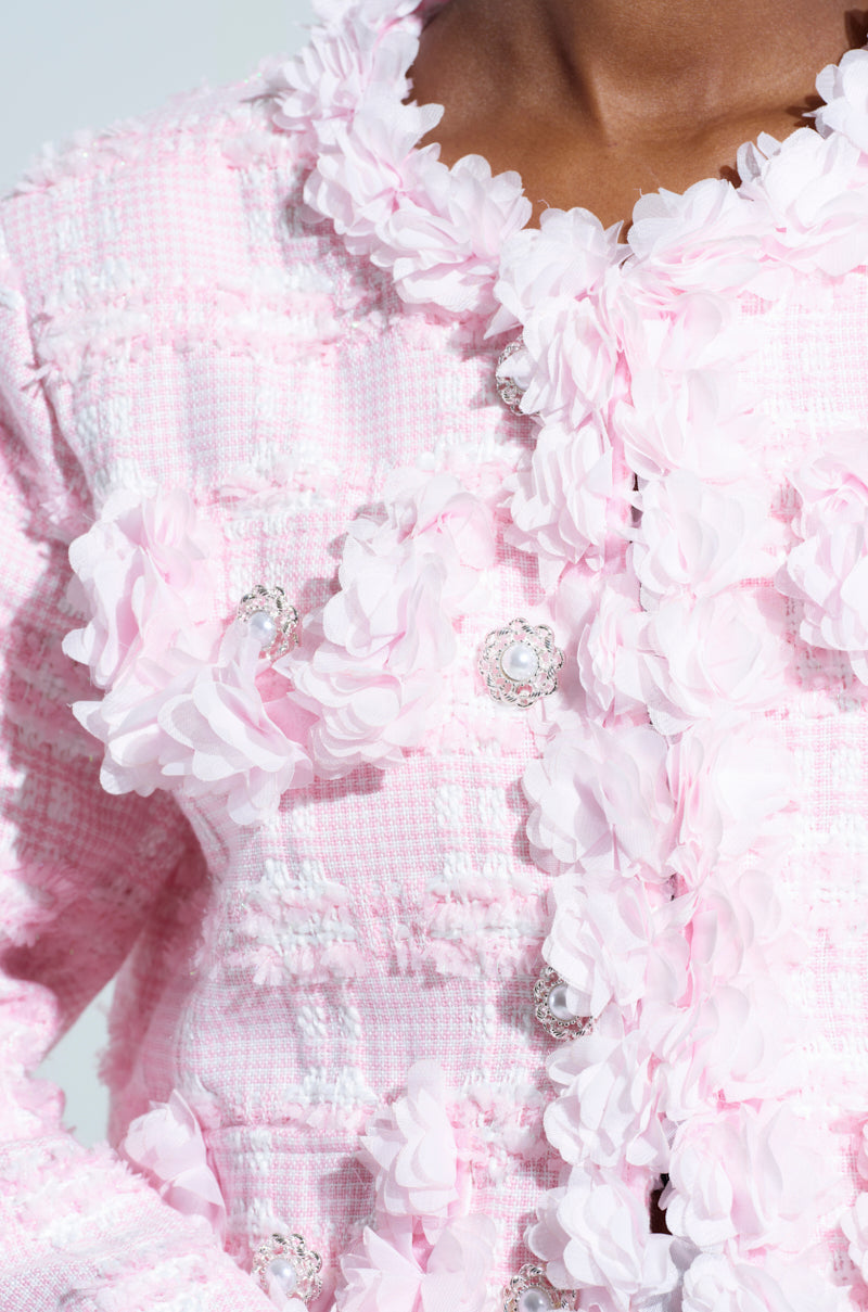 A close-up of the SPRINGTIME TWEED BLAZER highlights its light pink fabric embellished with delicate floral appliques. The decorative buttons add to its elegance, and the woven texture brings a charming touch to this sophisticated piece.